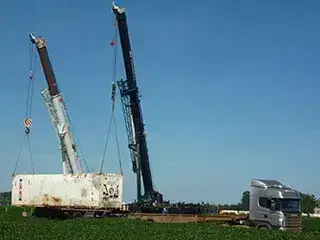 Guindastes levantam carga próxima a caminhão cinza.