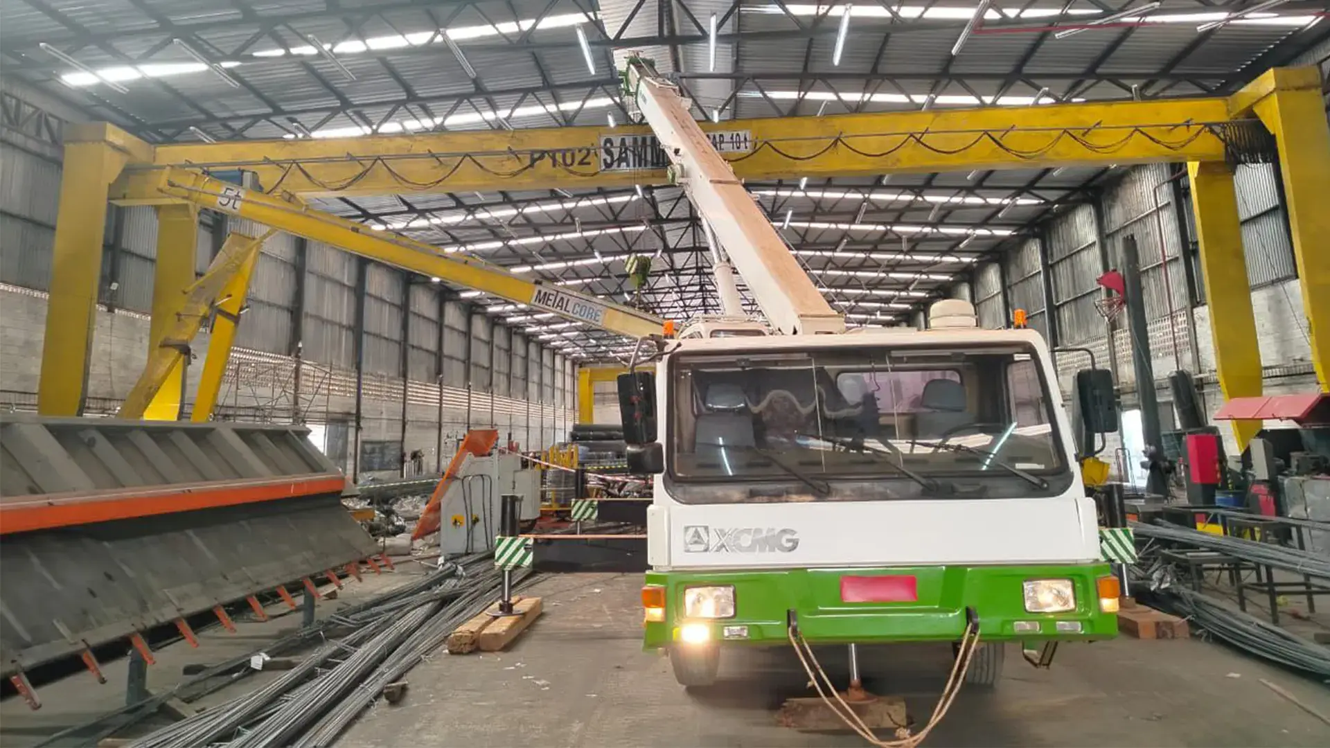 Caminhão guindaste XCMG realizando o içamento de uma ponte rolante amarela dentro de um galpão industrial