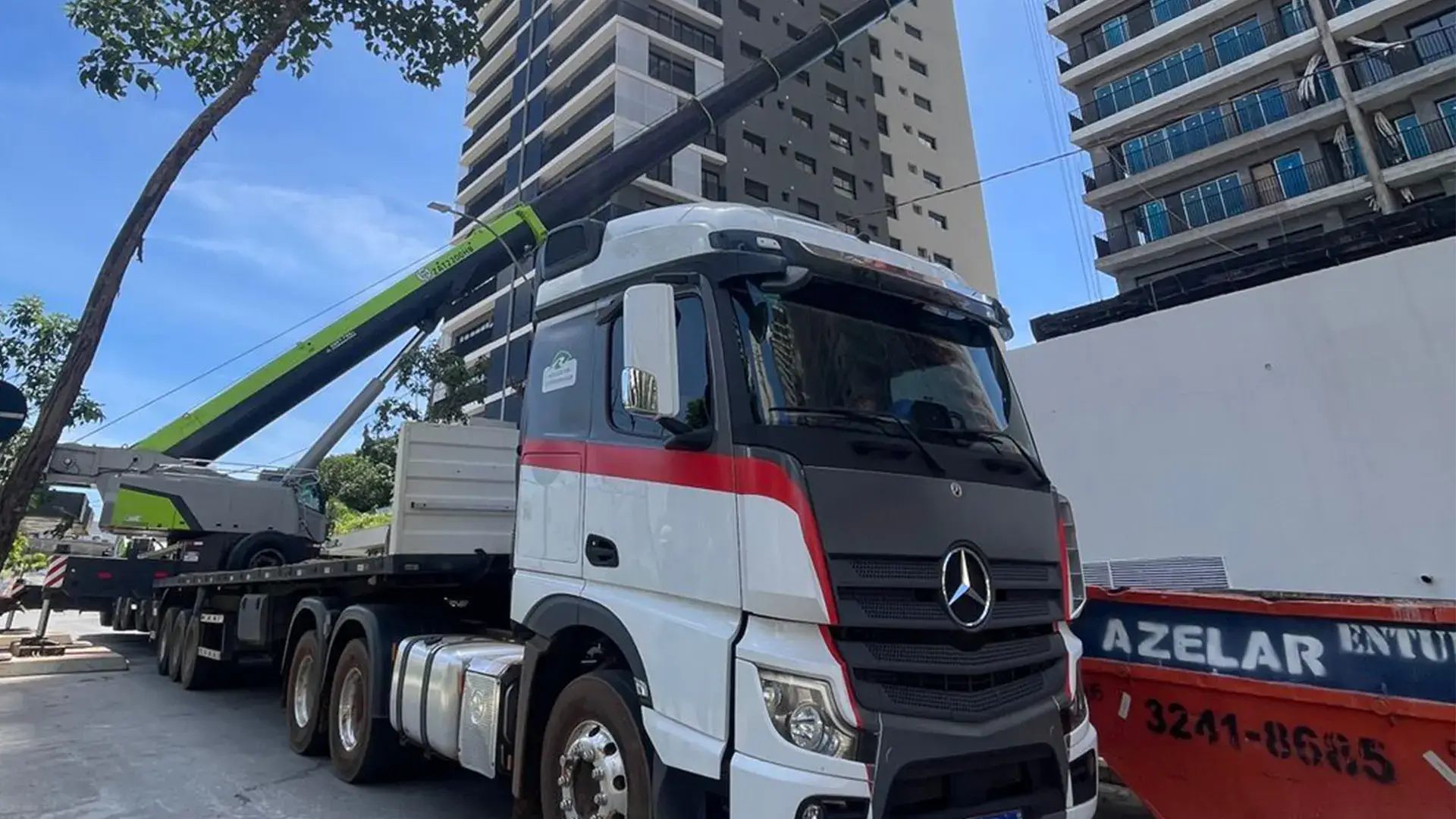 Caminhão Mercedes-Benz com guindaste telescópico verde realizando operação de içamento em grandes alturas próximo a edifícios residenciais.