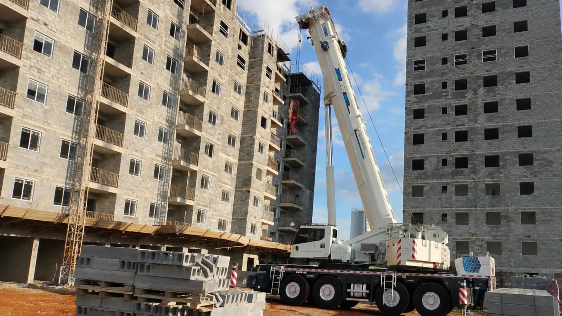 Guindaste móvel XCMG branco posicionado com estabilizadores em um canteiro de obras, operando entre dois grandes edifícios residenciais em fase de alvenaria estrutural.
