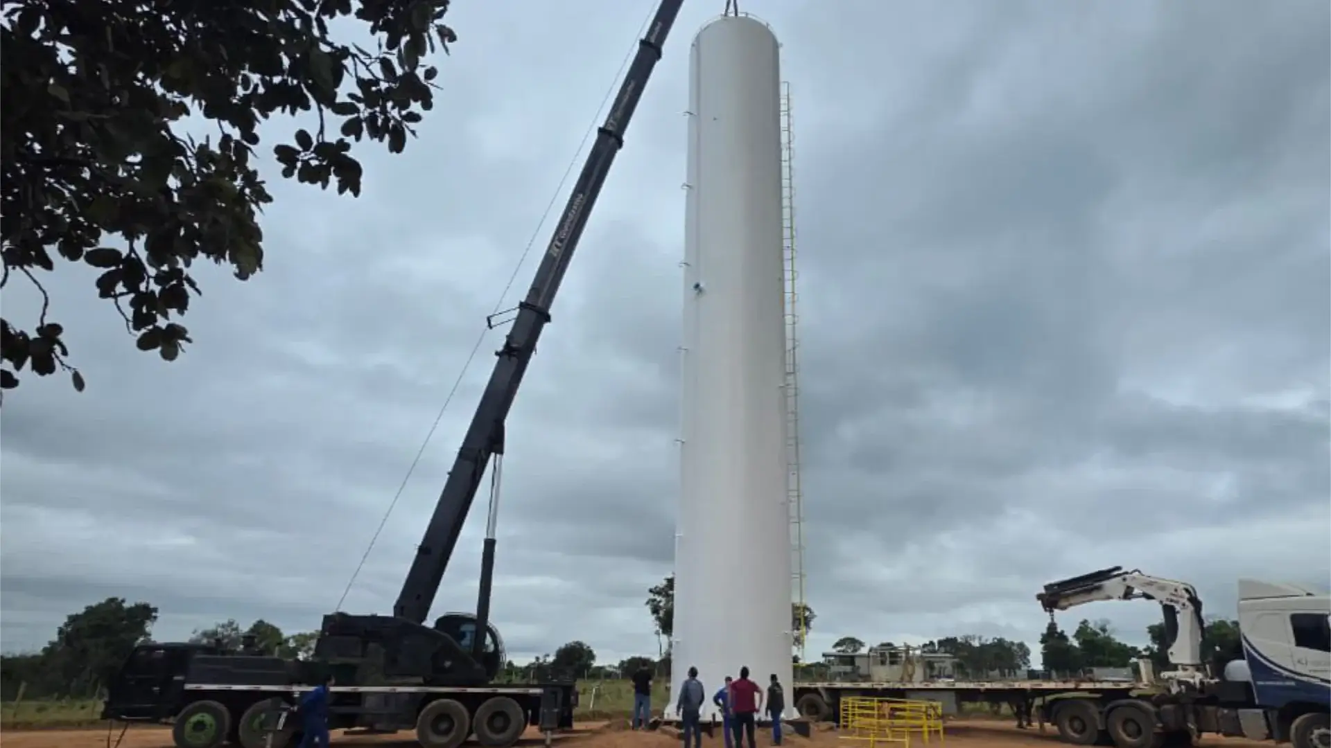 Guindaste preto realizando o içamento e a instalação de um grande reservatório de água cilíndrico e branco. Apoiados no solo, trabalhadores acompanham o posicionamento da estrutura ao lado de um caminhão de transporte em um canteiro de obras.